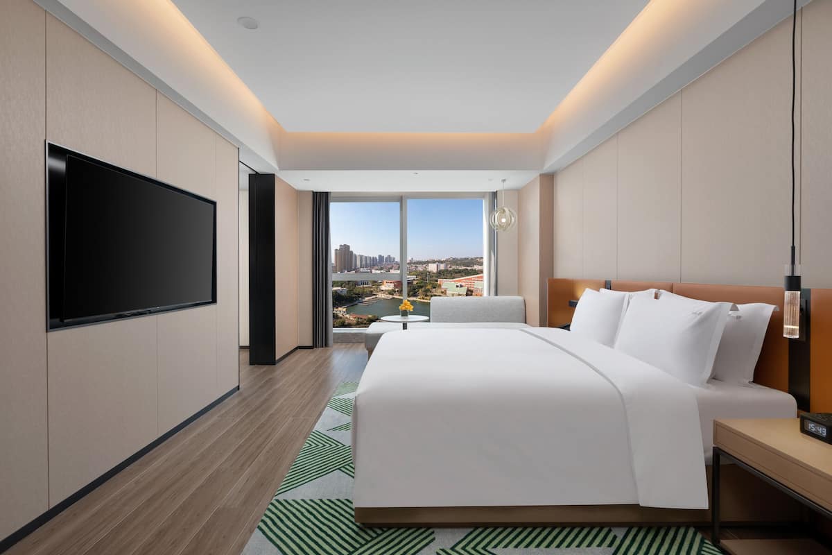 Una habitación de hotel moderna con una cama grande, un televisor de pantalla plana, una mesita y vistas a un paisaje urbano a través de amplios ventanales.