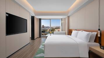 Una habitación de hotel moderna con una cama grande, un televisor de pantalla plana, una mesita y vistas a un paisaje urbano a través de amplios ventanales.