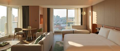 A hotel room with a large bed, a sofa, a dining table, and a view of the city.