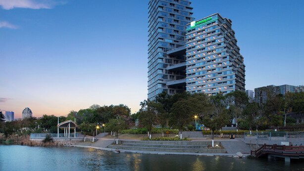 Een modern hoog hotel met een uniek hoekig ontwerp, gelegen nabij een waterkant met een park en een klein paviljoen.
