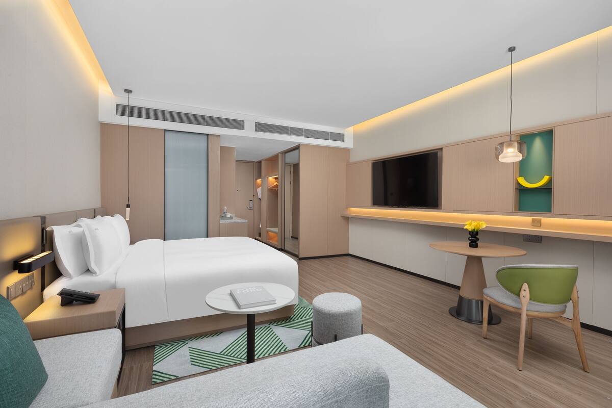Una habitación de hotel moderna con una cama, una mesita redonda, una silla verde, un sofá y un televisor montado en la pared.