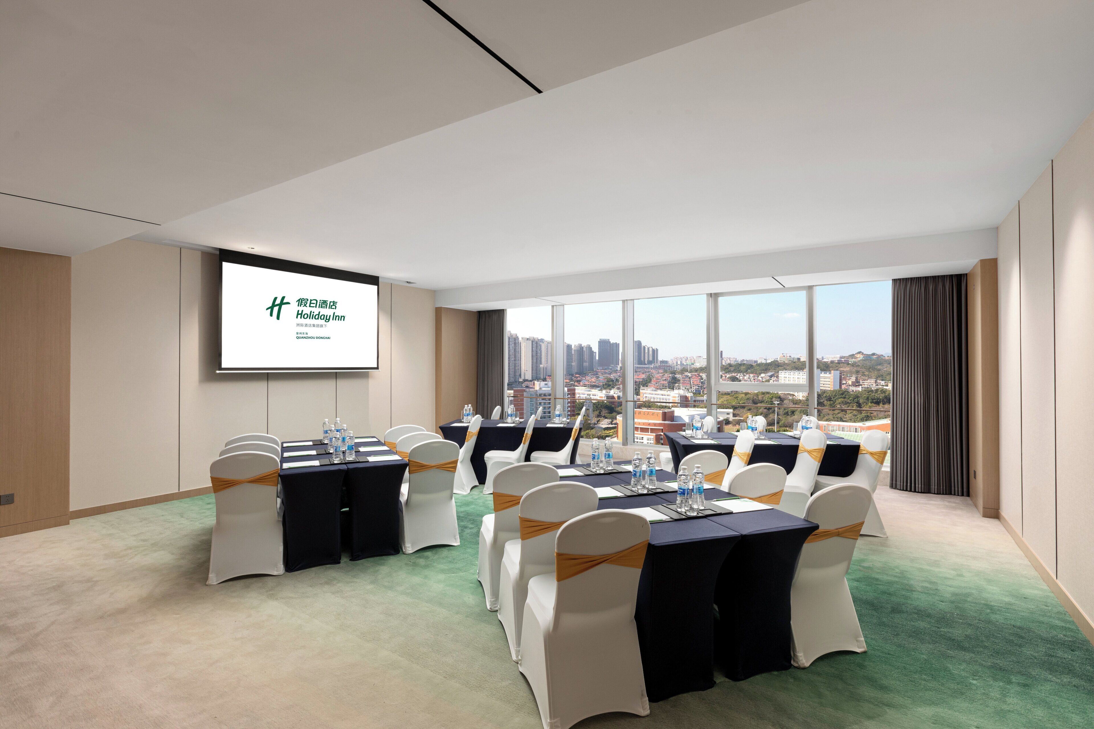 Un salón de conferencias con una pantalla grande mostrando una presentación, varias mesas con sillas y vistas a la ciudad a través de amplios ventanales.