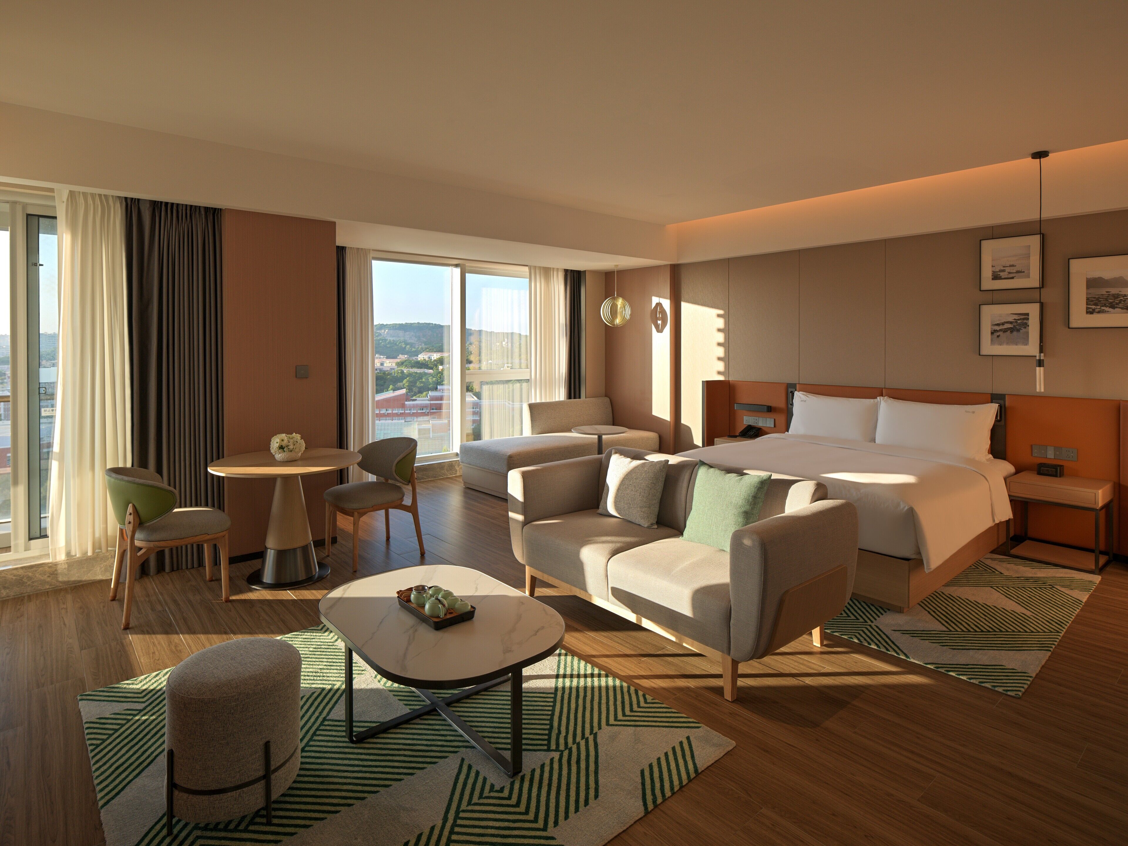 A modern hotel room with a large bed, two armchairs, a small table, and a view of the city.