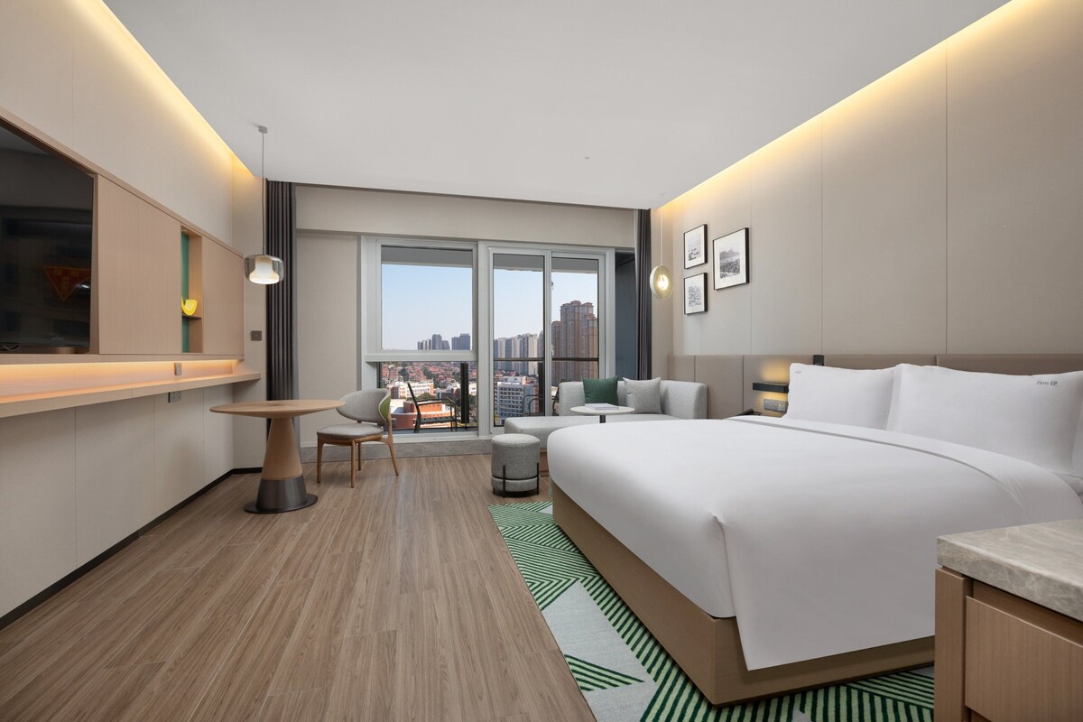 Un hotel moderno con una cama grande, un área de descanso con una mesa y sillas, vistas de la ciudad a través de amplias ventanas y un paisaje urbano visible.