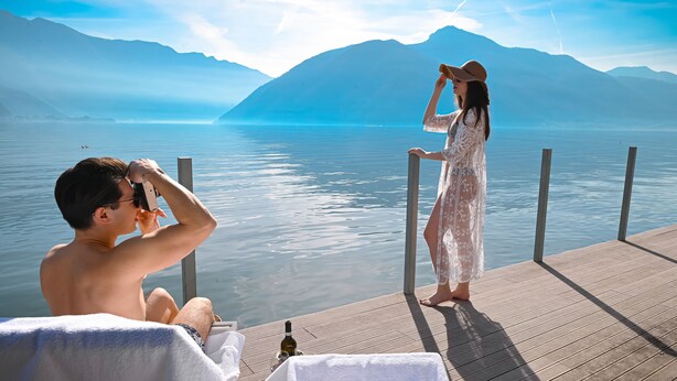 A couple on a wooden deck overlooking a calm lake with mountains in the background. - Lakeview Motta Penthouse in Melide (Melide)