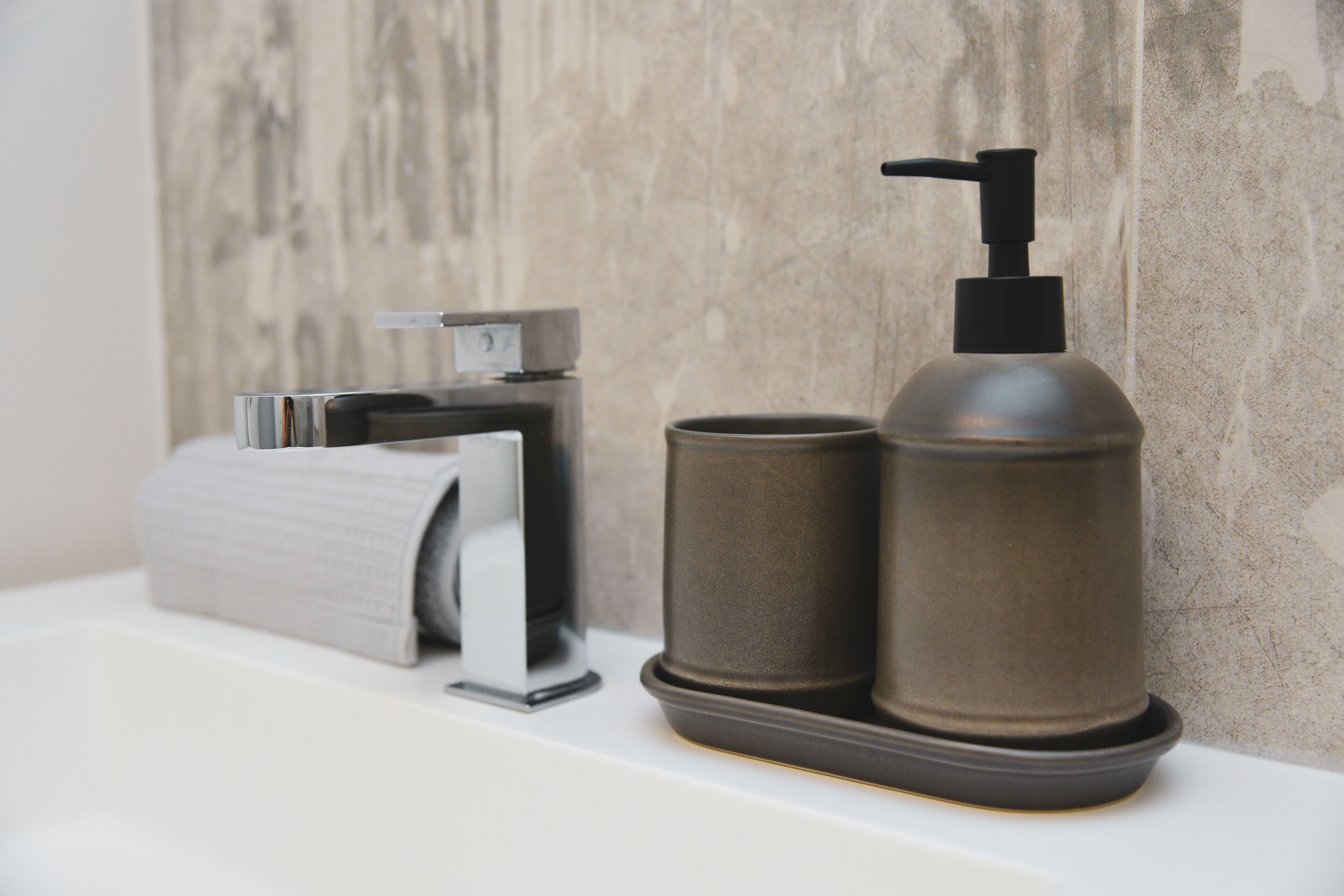 Une salle de bain avec un porte-rouleau de papier toilette, un distributeur de savon et un gobelet posé sur une surface blanche, le tout sur un mur à texture.