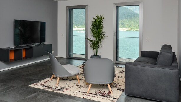 Una sala de estar moderna con un televisor de pantalla plana, dos sillones grises, un sofá gris y un ventanal con vistas a un lago y montañas.