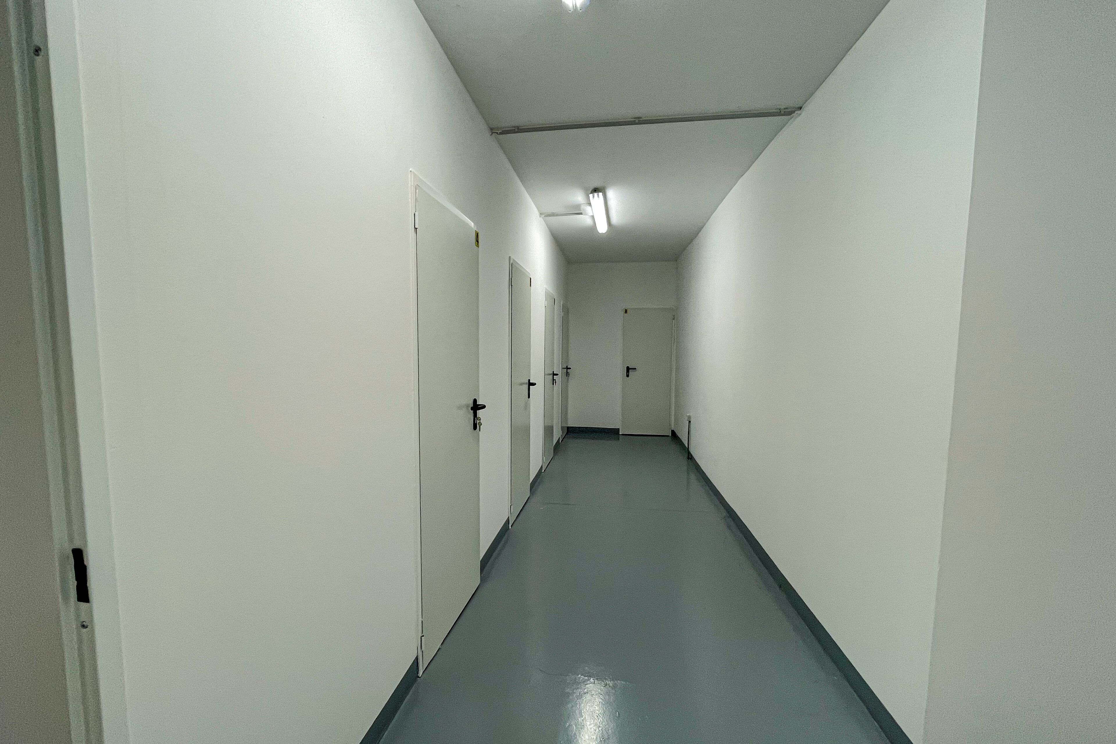 Un long couloir étroit aux murs blancs et au sol gris, qui mène à plusieurs portes.