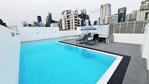 Une piscine sur le toit offrant une vue sur la ville, des chaises longues et un espace salon couvert.