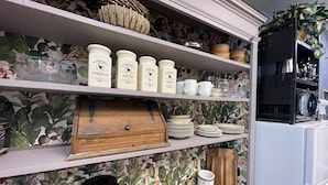 A kitchen with white shelves holding jars labeled 'Country Coffee,' 'Country Tea,' and 'Country Sugar.' There are also stacks of white plates and a wooden cutting board.