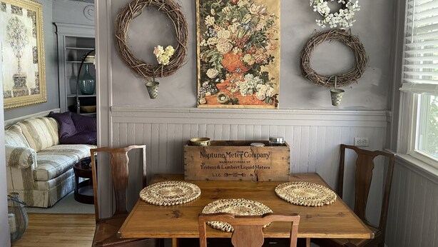 A dining area with a wooden table, four chairs, and a floral painting above.