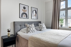 Un lit bien fait, recouvert d’une courtepointe matelassée, avec une tête de lit rembourrée de couleur grise et des oreillers à motifs. Deux œuvres d’art abstraites encadrées sont accrochées au mur au-dessus du lit.