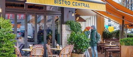 An outdoor cafe named Bistro Central with wicker chairs and tables, a large planter, and a yellow awning.