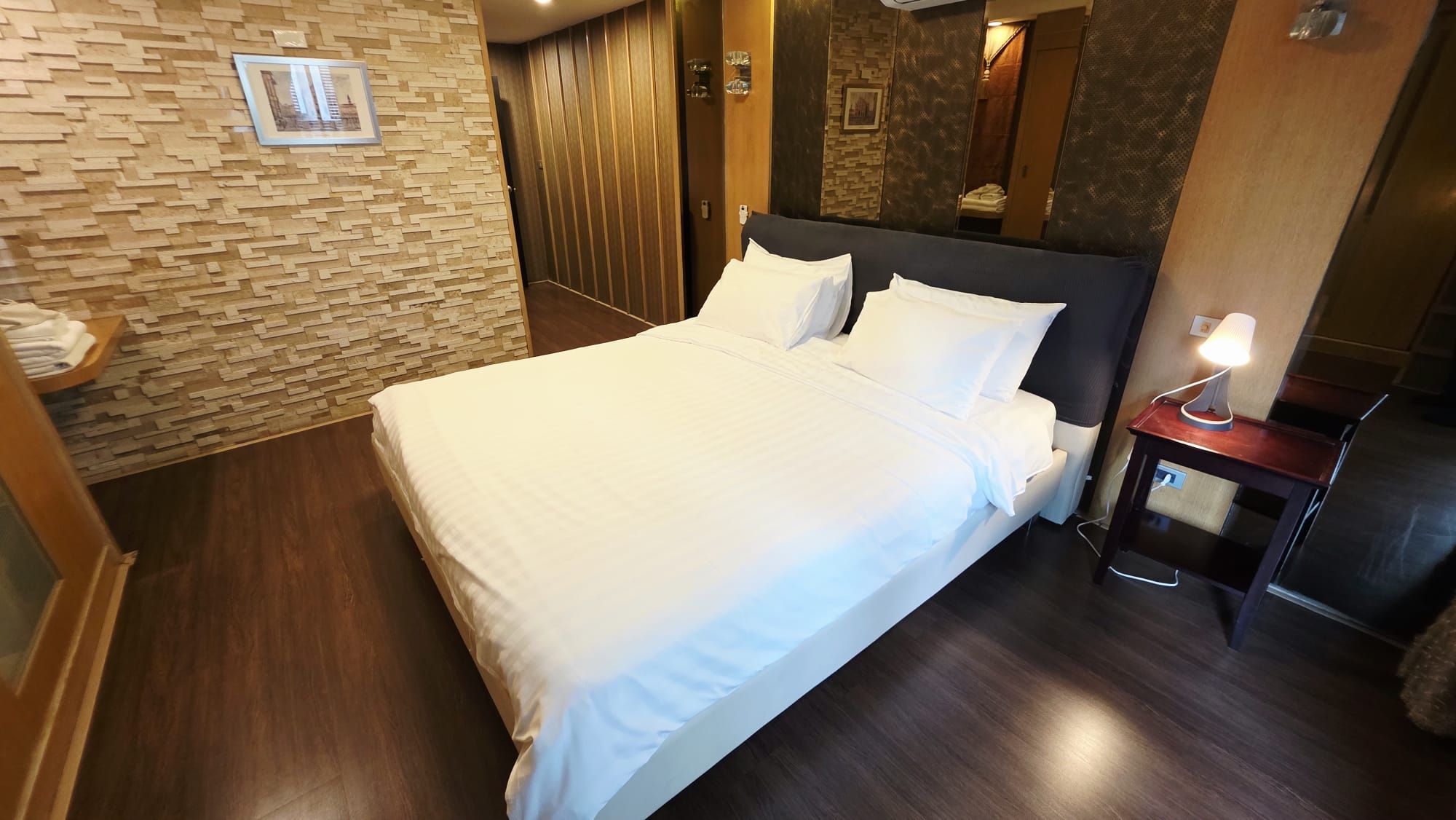 A hotel room with a bed, a bedside table with a lamp, a wooden floor, and a wall with a textured pattern.