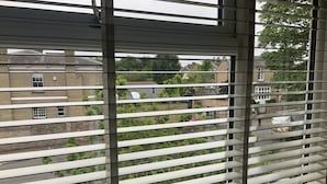 Uitzicht door jaloezieën, waarop een bakstenen gebouw met meerdere ramen en een groen landschap met bomen zichtbaar is.