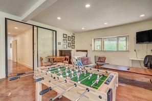 A room with a foosball table, a sofa, a television, and a window with a view of greenery.