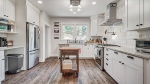 A modern kitchen with white cabinetry, a stainless steel refrigerator, a wooden cart, and a window providing natural light. - 3BD Cottage Backyard near Garden of the Gods in CO (Colorado Springs)