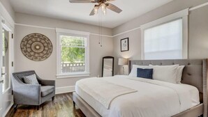 A bedroom with a large bed, a ceiling fan, a window with a view of greenery, a chair, and a decorative wall piece. - 3BD Cottage Backyard near Garden of the Gods in CO (Colorado Springs)