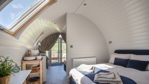 A bedroom with a skylight, a bed with a blue blanket, a wooden table with kitchenware, and a view of the outdoors. - Cedar (Bridport)