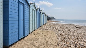 A row of blue beach huts along a pebbly beach with the sea in the background. - Cedar (Bridport)