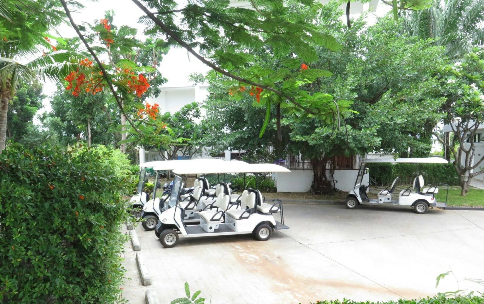 A golf cart with passengers, parked on a concrete surface with trees and greenery in the background.