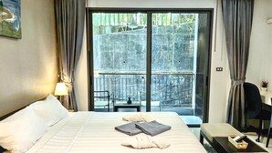 Habitación de hotel con cama, escritorio, silla, balcón con vistas a árboles y un cuadro en la pared.