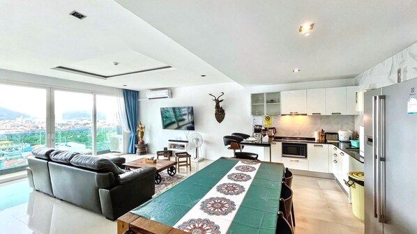 A modern living room with a sofa, a dining table with a patterned runner, a flat-screen TV, and a kitchen area with cabinets and appliances. - Luxurious 4-Bedroom Apartment with Stunning Patong Bay Views (Pa Tong)
