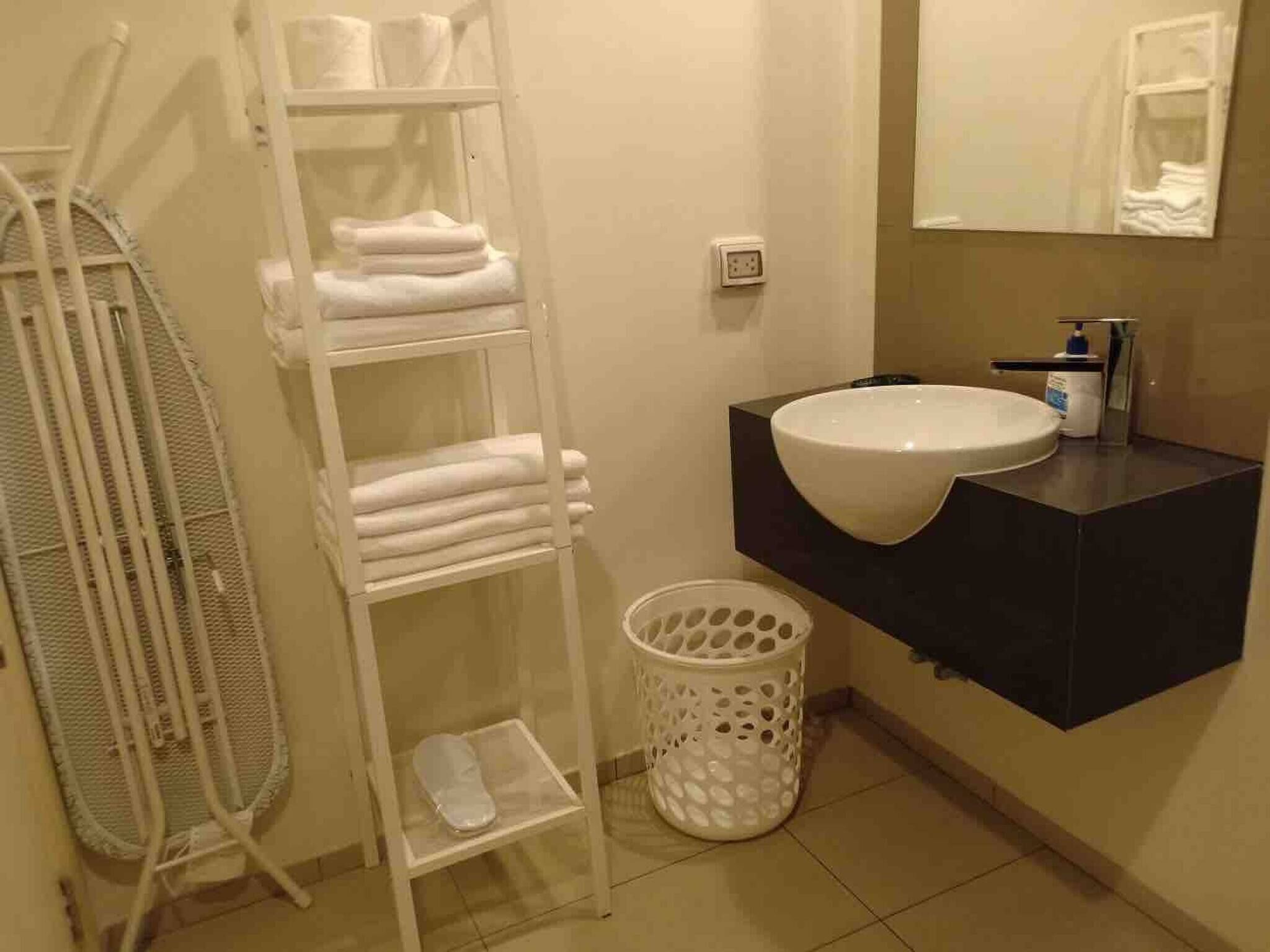 Ein Badezimmer mit einem weißen Regal mit Handtüchern, einem weißen Wäschekorb, einem weißen Waschbecken mit dunkler Arbeitsplatte und einem Spiegel über dem Waschbecken.
