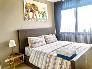 A hotel room with a bed, a nightstand, a lamp, a painting of elephants, and a window with curtains.