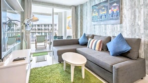Ein modernes Wohnzimmer mit einem grauen Sofa, einem kleinen weißen Couchtisch und einem grünen Teppich. Ein großes Fenster bietet Blick auf Gebäude und einen Balkon.