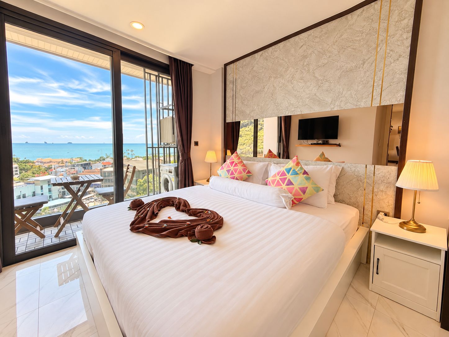 Habitación de hotel con una cama grande, televisión, balcón con vista al mar y una cama bien hecha con una toalla con forma de corazón.
