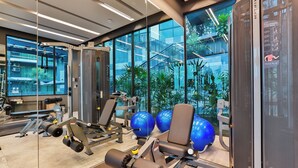 Een sportschool met trainingsapparatuur, grote ramen en uitzicht op een gebouw met trappen en groen.