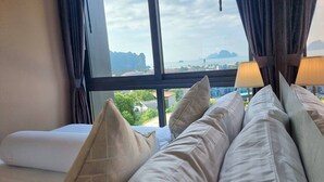 Een hotelkamer met een bed, kussens, een raam met gordijnen en uitzicht op zee en gebouwen.