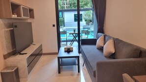 A modern living room with a flat-screen TV, a small table with a plant, a sofa with cushions, and a view of a swimming pool and outdoor area. - B207 - 1 Bedroom Serviced Apartment with Pool Access at Ao Nang Beach (Tambon Ao Nang)