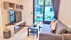 A modern living room with a flat-screen TV, a sofa, a coffee table, and a view of an outdoor swimming pool. - B207 - 1 Bedroom Serviced Apartment with Pool Access at Ao Nang Beach (Tambon Ao Nang)