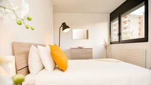 Une chambre moderne comprenant un lit, une table de chevet, une lampe, une commode, un miroir et une fenêtre donnant sur des immeubles.