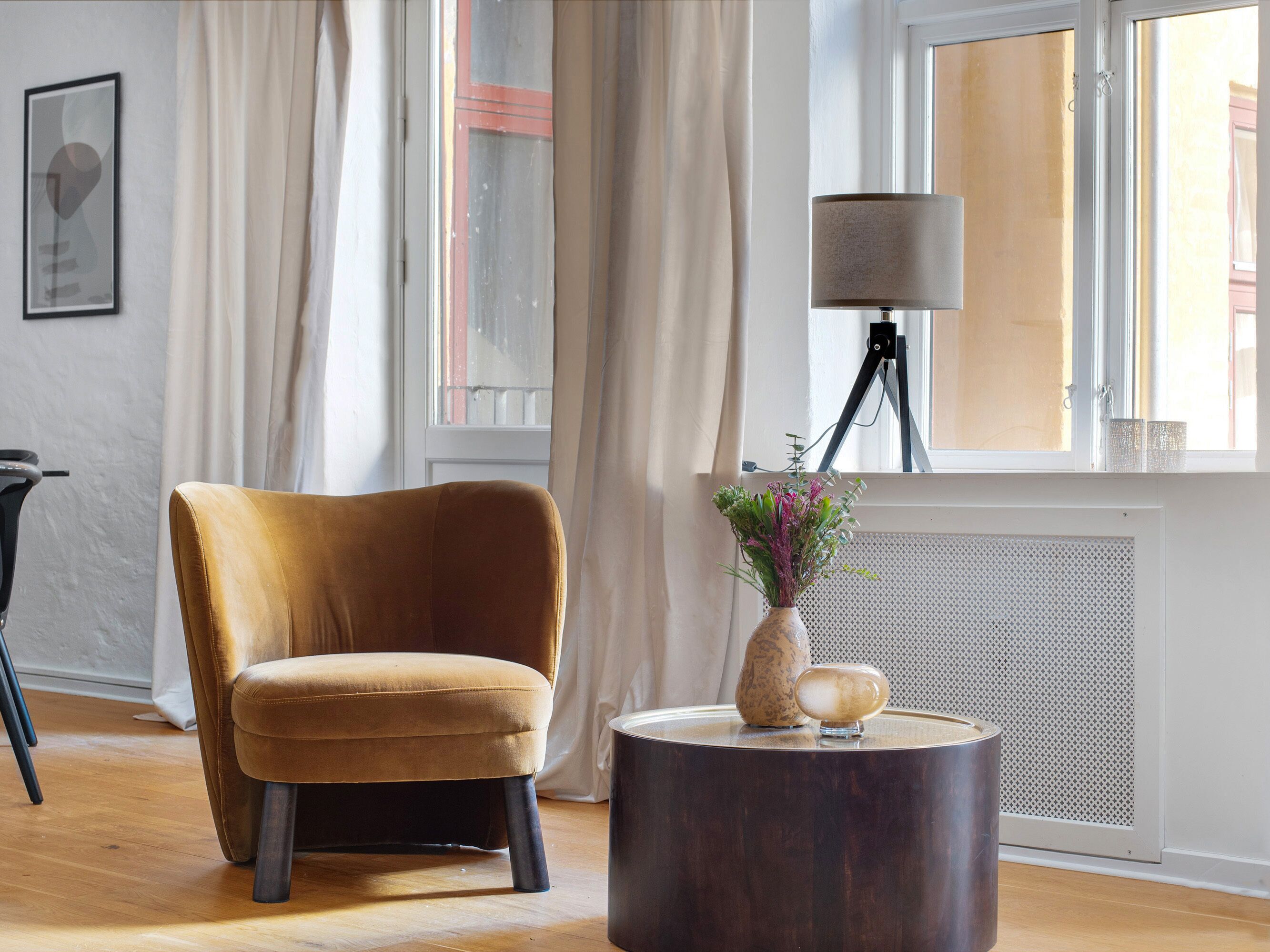 Een kamer met een mosterdkleurige fauteuil, een rond houten bijzettafeltje met een vaas en decoratieve objecten, een vloerlamp en een radiator.