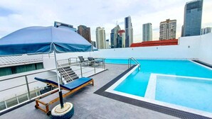 A rooftop pool with a city skyline in the background, a covered area with lounge chairs, and a staircase leading to an upper level.