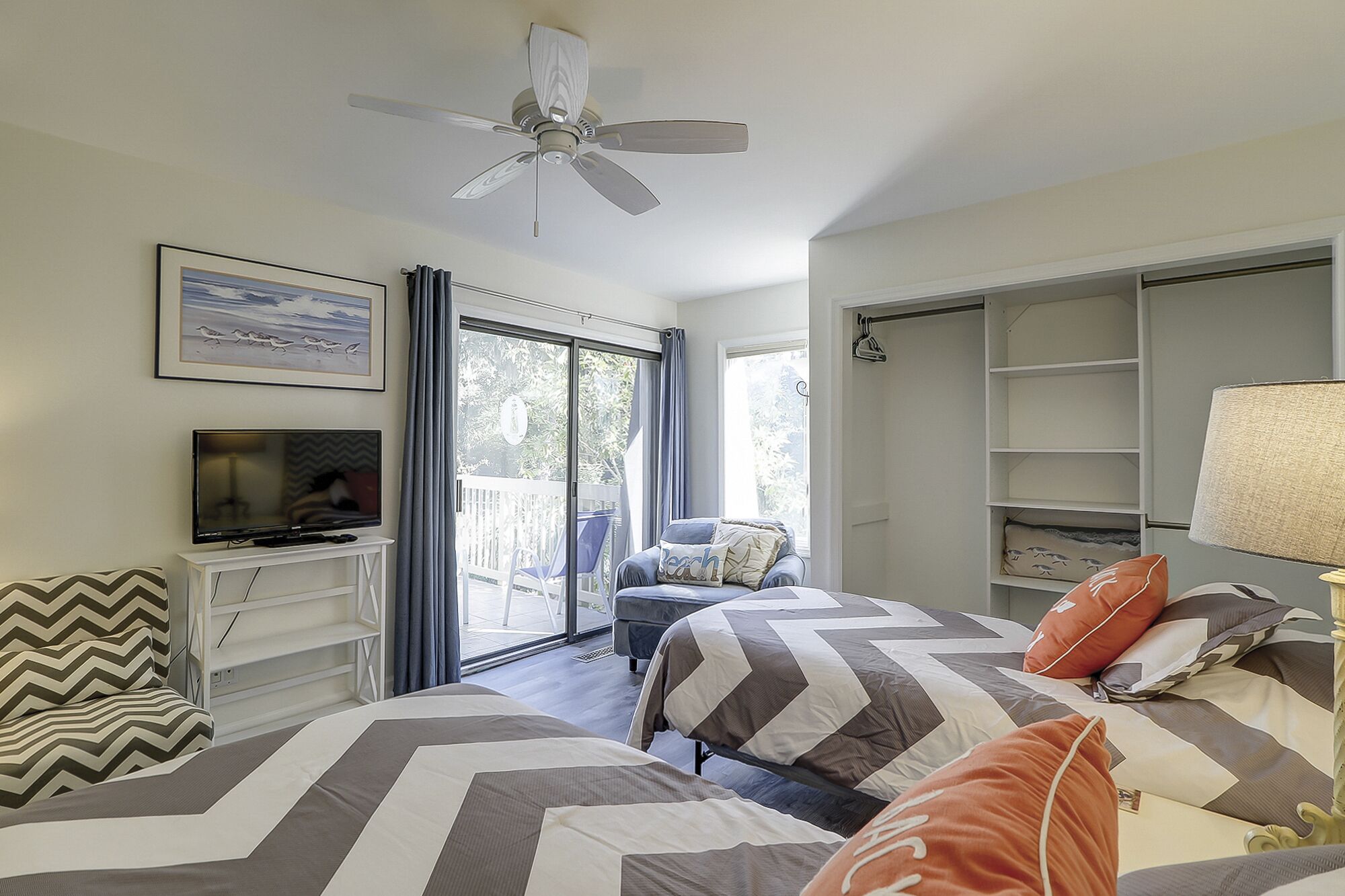 A bedroom with two beds, a ceiling fan, a TV, a chair, and a sliding glass door to a balcony.