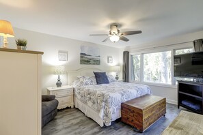 Un dormitorio con cama, mesitas de noche, ventilador de techo, un cofre de madera, televisor y ventana con cortinas.