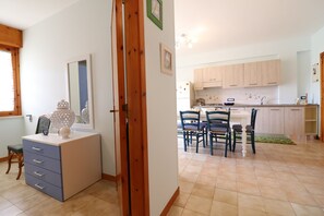 A room with a dresser, a chair, a dining table with chairs, a kitchen area with cabinets, and a mirror.