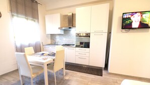 Una cocina moderna con armarios blancos, una estufa a gas y un horno empotrado. Hay una mesa de comedor con sillas y un televisor montado en la pared.
