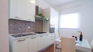 A modern kitchen with white cabinets, a stainless steel sink, and a built-in oven. There is a dining table with two chairs and a bottle of wine on it.