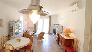 Uma sala de jantar com uma mesa e cadeiras brancas, um lustre pendurado e uma televisão sobre um móvel.