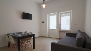 A living room with a sofa, a dining table with chairs, a flat-screen TV mounted on the wall, and a sliding glass door leading to a balcony.