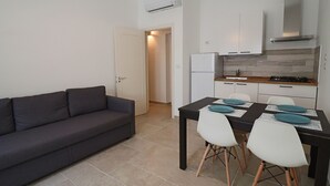 A compact living space with a dark gray sofa, a dining table with white chairs, and a kitchenette with a stove and refrigerator.