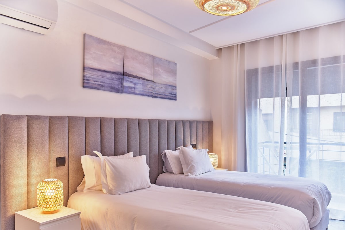 Ein modernes Schlafzimmer mit zwei Betten, einem großen Fenster mit durchsichtigen Vorhängen und einem an der Wand befestigten Kunstwerk.