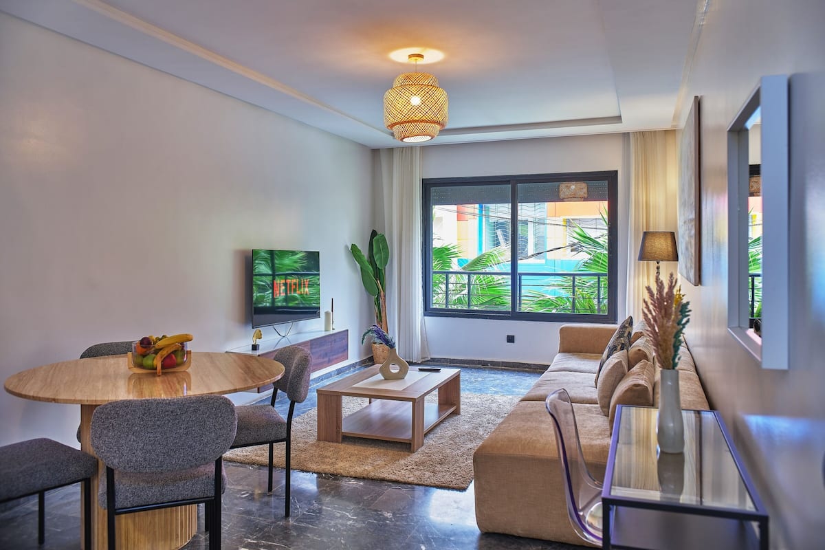 Ein modernes Wohnzimmer mit Fernseher, Essbereich, Sofa und großen Fenstern mit Blick auf Grünflächen.