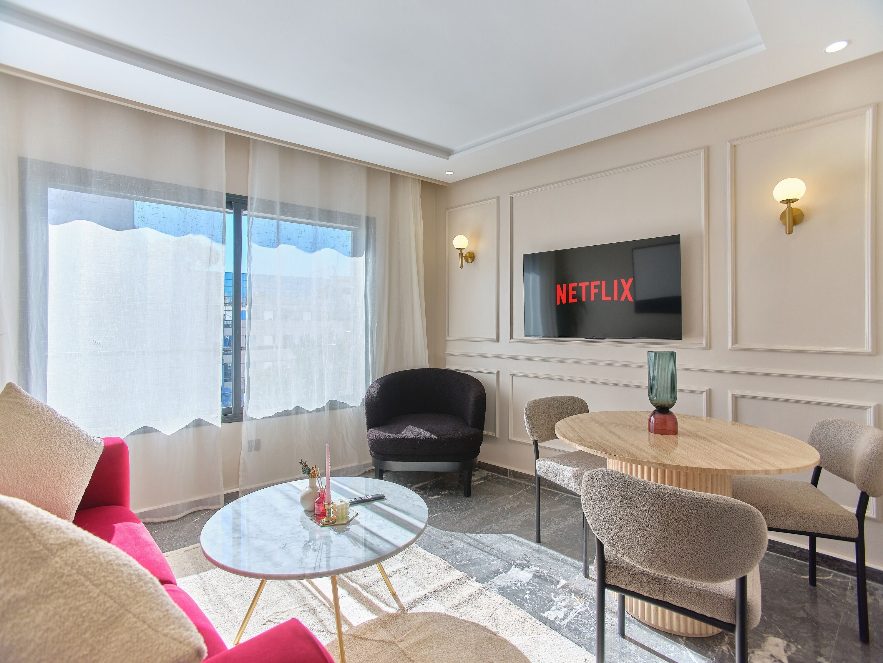A hotel room with a large window, a flat-screen TV mounted on the wall, a round glass table, and several chairs.