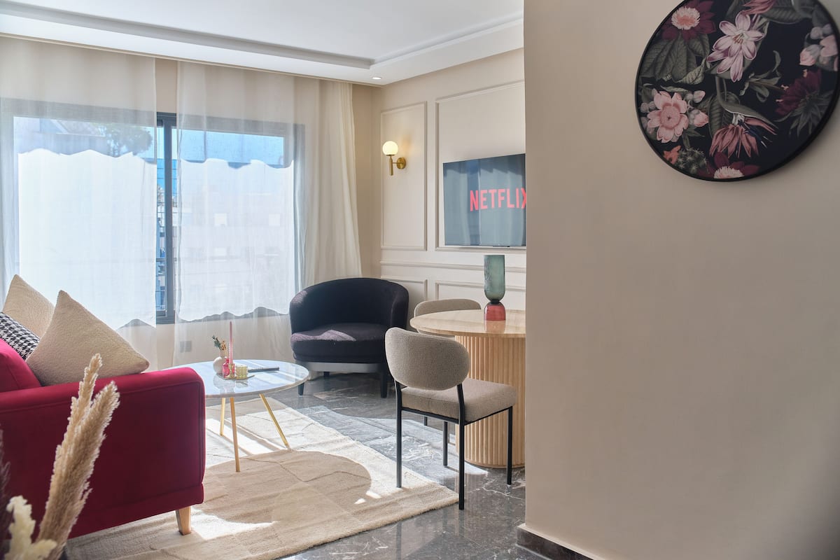 Ein modernes Wohnzimmer mit einem roten Sofa, einem schwarzen Sessel, einem kleinen runden Tisch und einem Couchtisch. Ein großes Fenster mit durchsichtigen Vorhängen lässt natürliches Licht herein.
